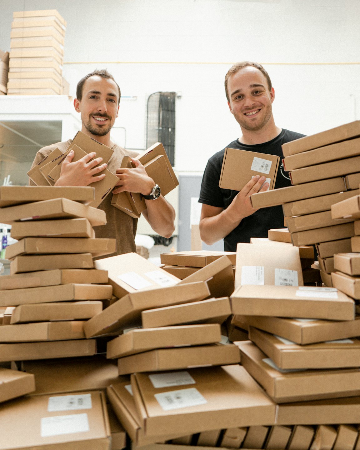 Two men smiling among shipping boxes in a warehouse.